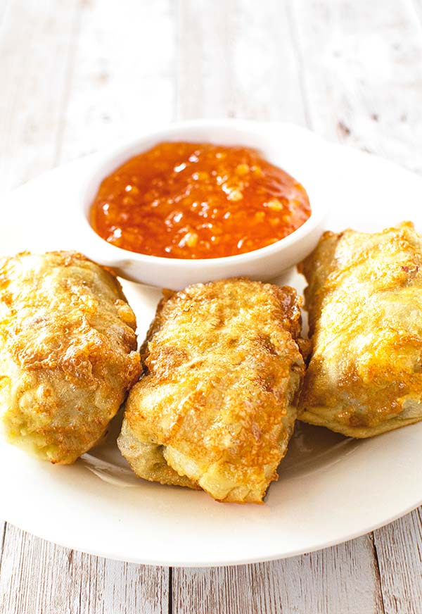 Three egg rolls on a white plate with a small white bowl of duck sauce on a white wooden table