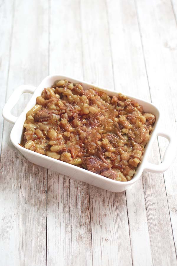 Thick cholent in a white trangular serving dish on a white wood table.