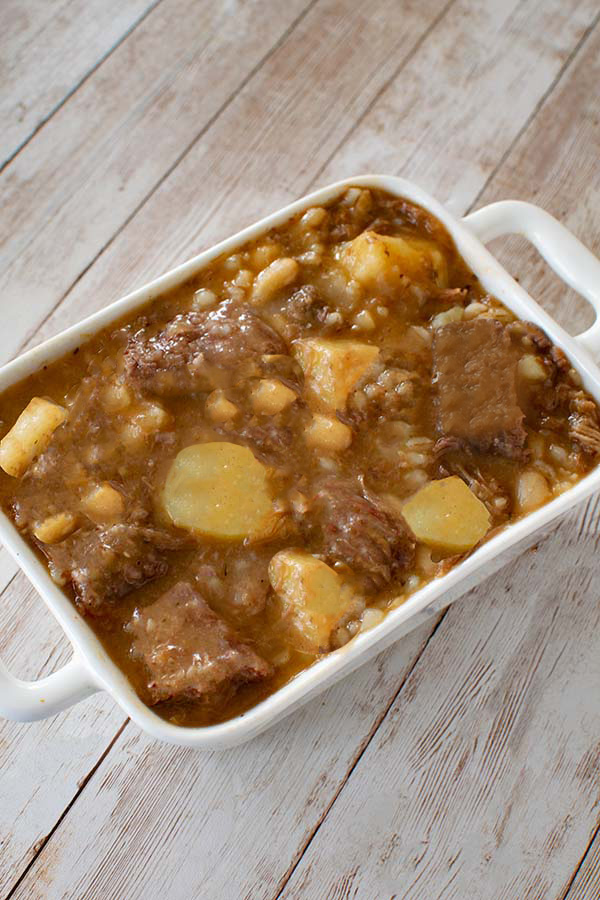 Cholent in a white rectagular ceramic serving dish on a white plank board backgorund