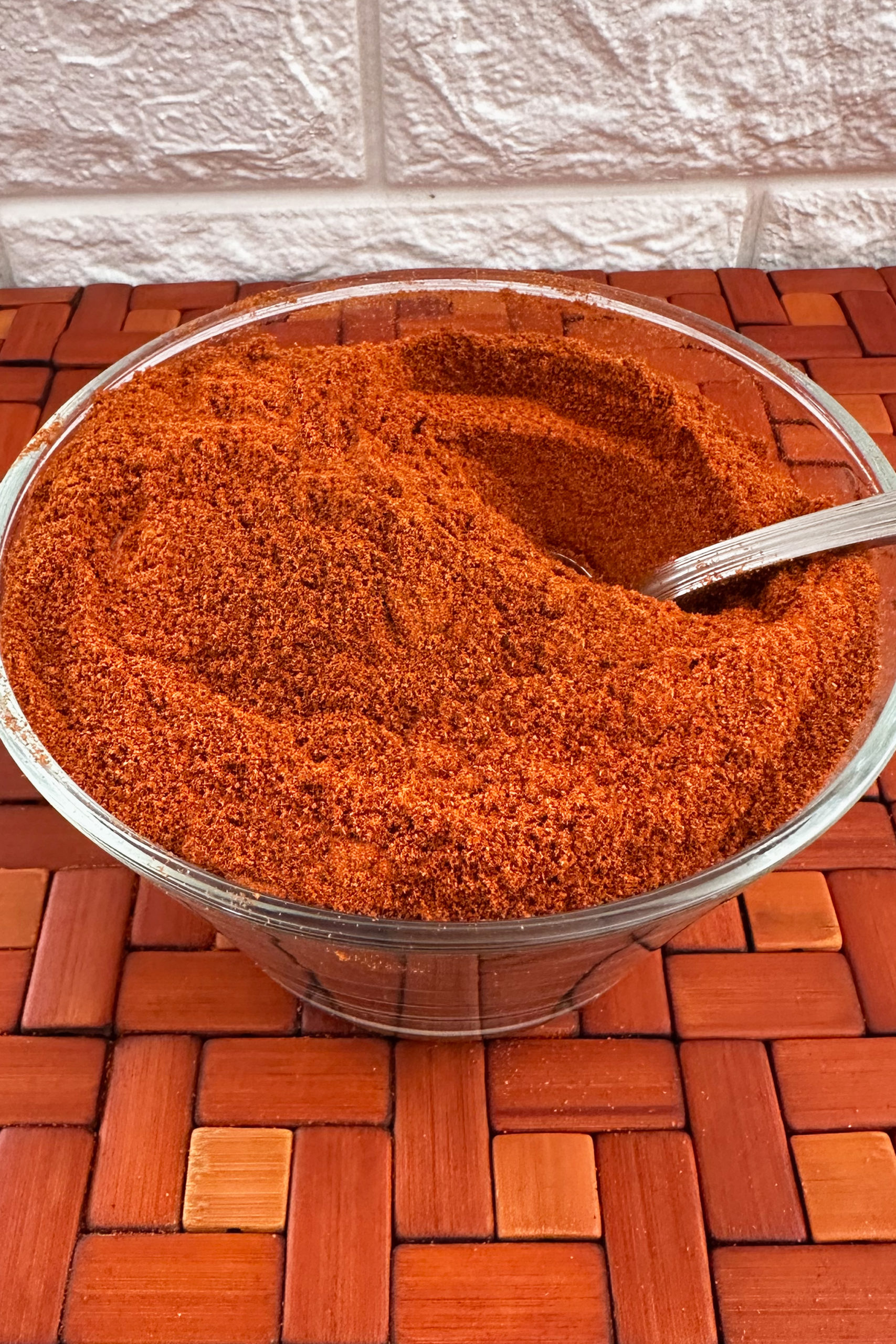 Israeli Grill Spice and BBQ Seasoning in a clear bowl with a spoon on a wooden mat.