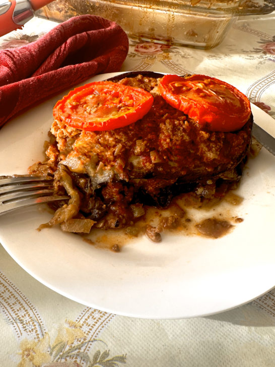 Israeli-style moussaka with tomato slices on top and a fork and a knife, all on a white plate on a cream colored table cloth with flowers and a red cloth napkin nearby. In the