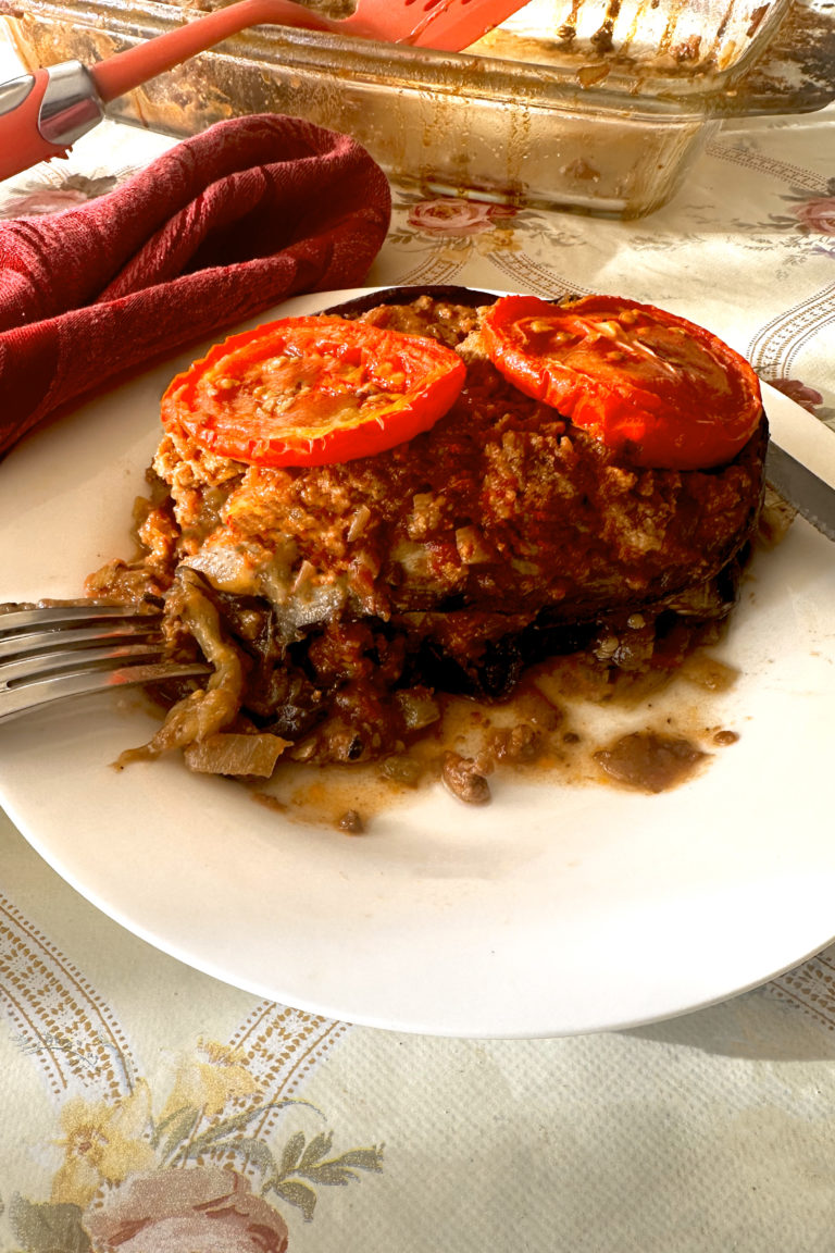 Israeli-style moussaka with tomato slices on top and a fork and a knife, all on a white plate on a cream colored table cloth with flowers and a red cloth napkin nearby. In the