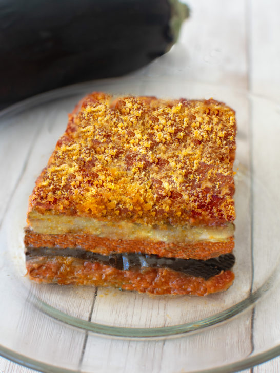 Slice of moussaka on a clear plate on a white wood table.