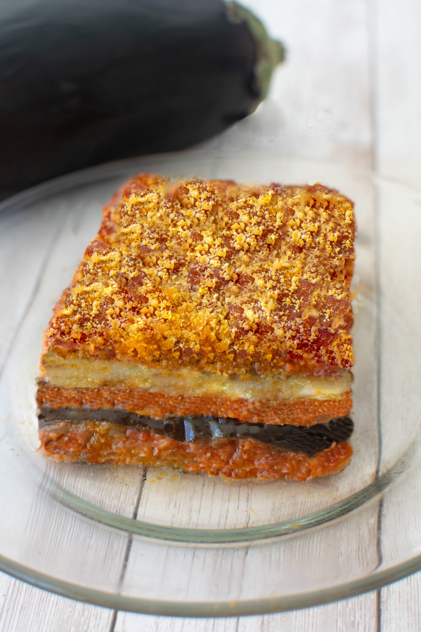 Slice of moussaka on a clear plate on a white wood table.