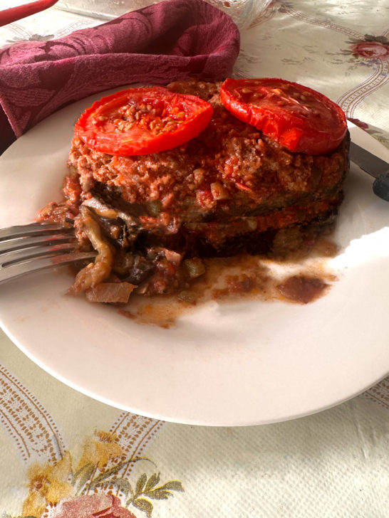 A large slice of Israeli-Style Moussaka with slices of tomato on top with a fork and an knife on a white plate