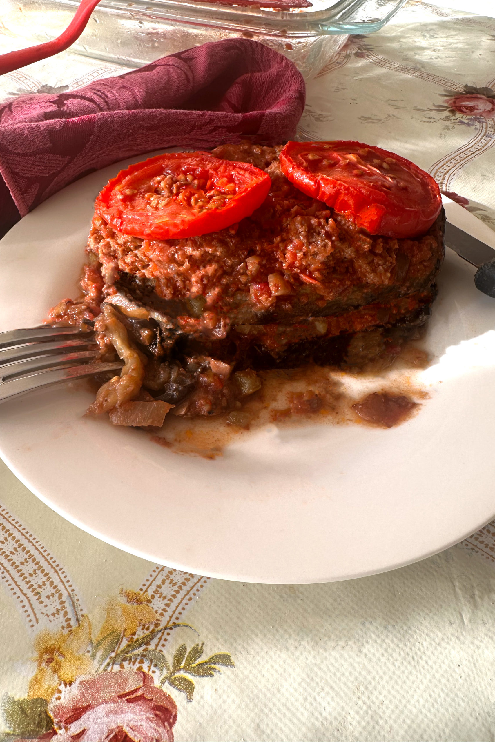 A large slice of Israeli-Style Moussaka with slices of tomato on top with a fork and an knife on a white plate next to a red cloth napkin with a glass serving dish in the background on a tablecloth with flowers