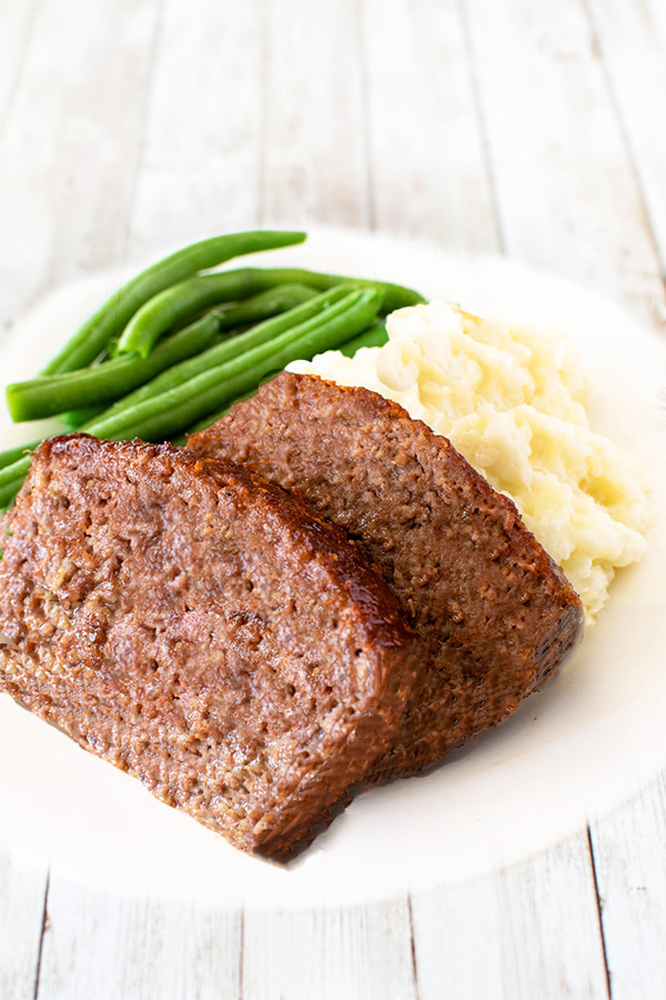 2 slices of meatloaf on a white plate with mashed potatoes and long green beans