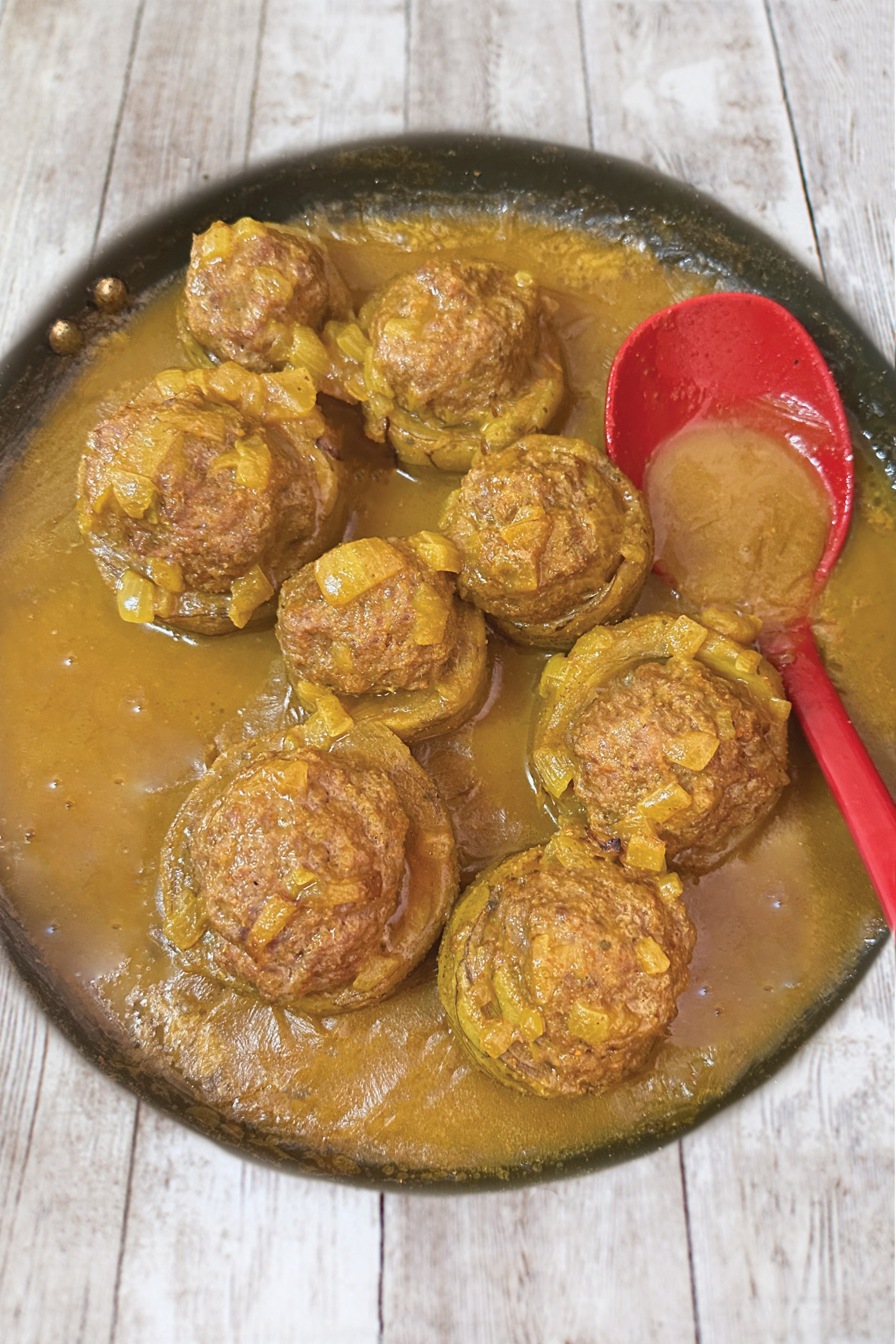 Israeli-Style Stuffed Artichoke Bottoms in cumin sauce with a red spoon on a white wood table.