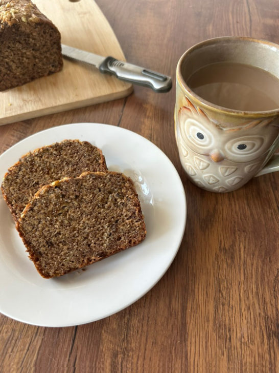 Slices of Dairy-Free Banana bread near a cutting board with the banana loaf on it with a knife and near an owl mug of coffee, all on a dark wood table.