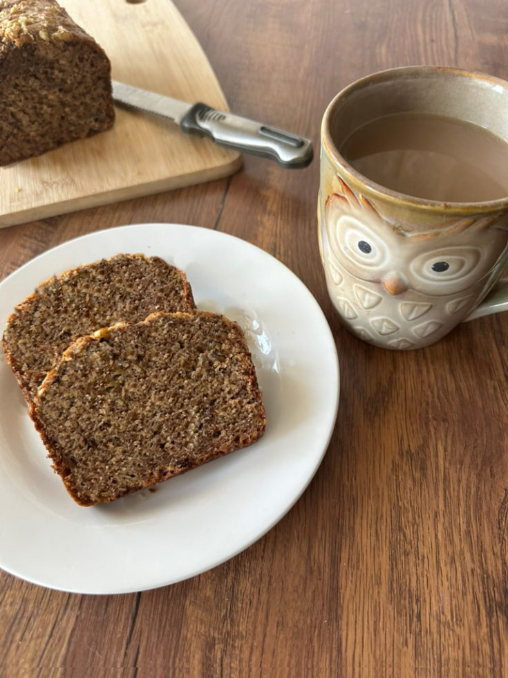 Slices of Dairy-Free Banana bread near a cutting board with the banana loaf on it with a knife and near an owl mug of coffee, all on a dark wood table.