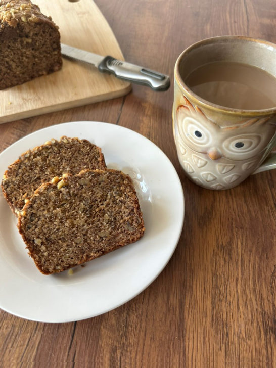 Slices of easy Banana bread with walnuts near a cutting board with the banana loaf on it with a knife and near an owl mug of coffee, all on a dark wood table.