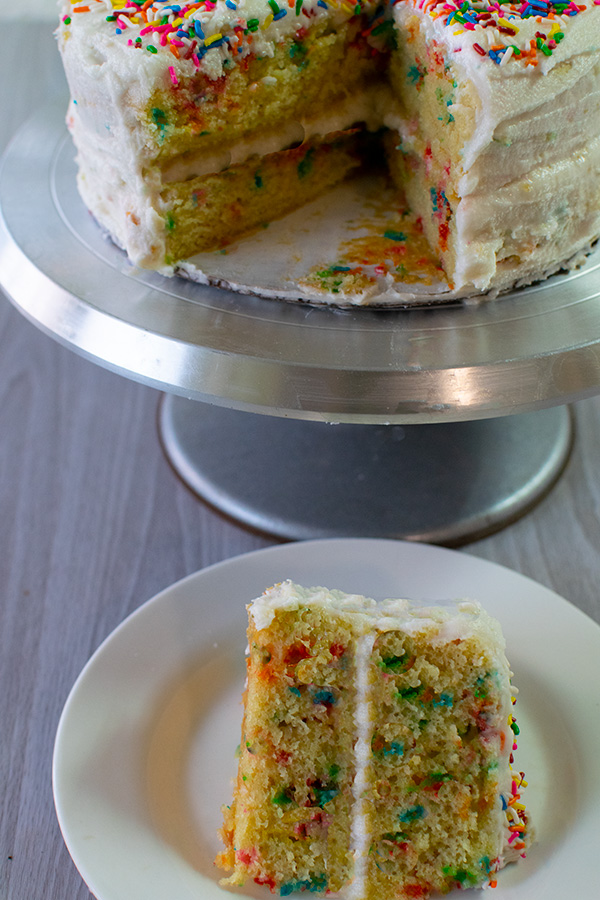 Dairy Free Funfetti Cake on a stand with a slice missing and the slice on a white plate in front - all on a white wood background