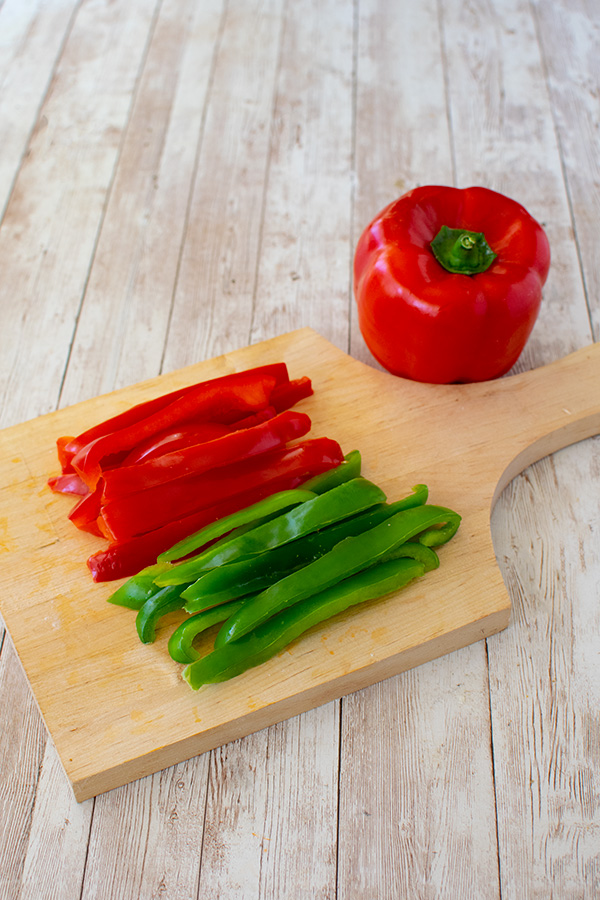 Red and green pepper slices on a wooden cutting board with a red pepper nearby all on a white wood background