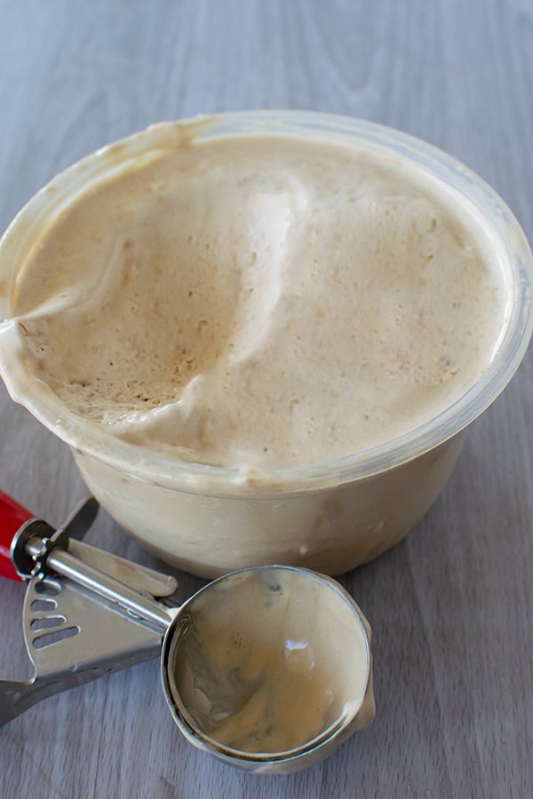 A small tub of coffee ice cream missing a scoop of ice cream and a used ice cream scoop on a white wood background