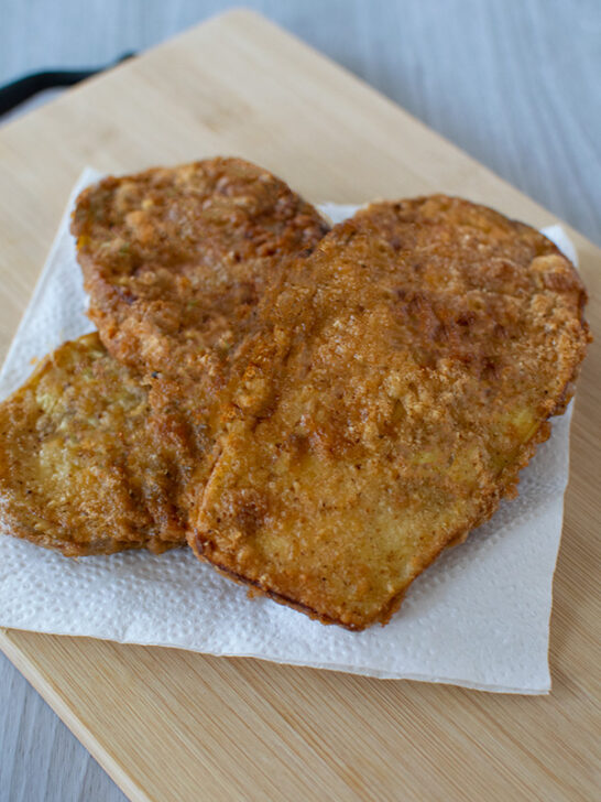 Three pieces of crispy, golden brown slices of eggplant (eggplant cutlets) ona white napkin on a wooden cutting board.