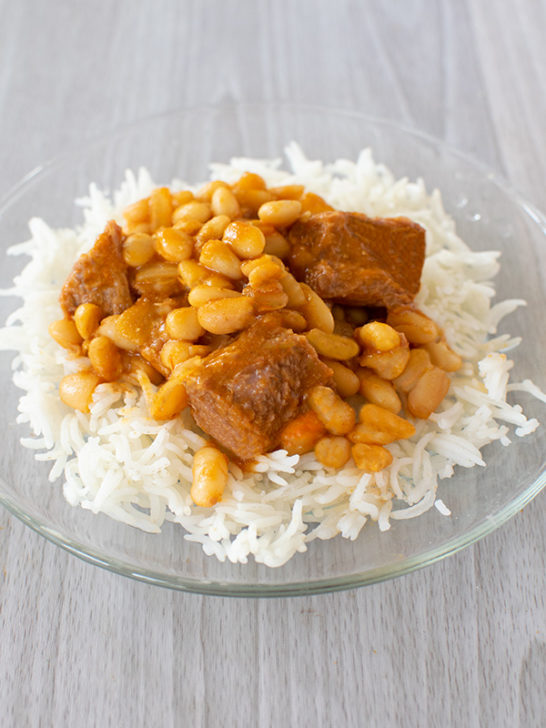 Israeli bean soup with chunks of meat over rice on a clear plate and all on a white wood background