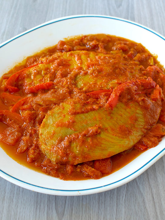 Two pieces of Moroccan fish with peppers on a white plate with a blue line around the edges on a white wood background