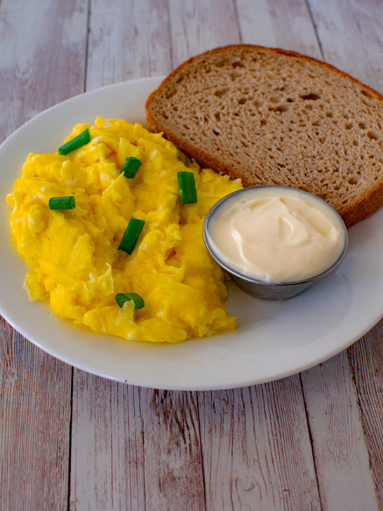 Diary free scrambled eggs on a white plate with a piece of matzo and a little metal bowl of mayonnaise on a white wood table.