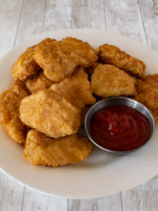 Cornflake dairy free chicken nuggets with a little metal bowl on a white plate on a white wood table