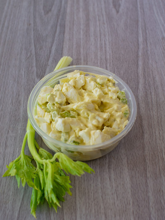 Deli egg salad in a container next to a celery stick on a gray wood table.