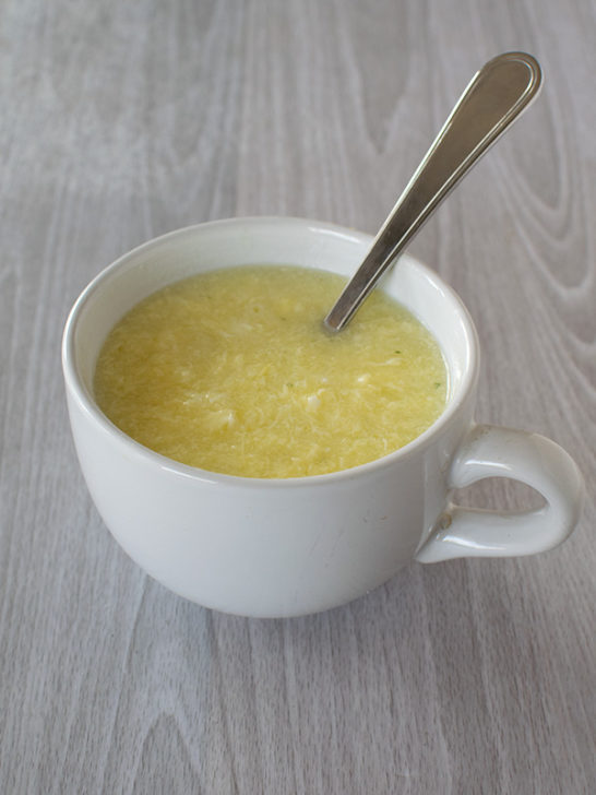 Egg drop soup in a white soup mug with a spoon on a white wood table