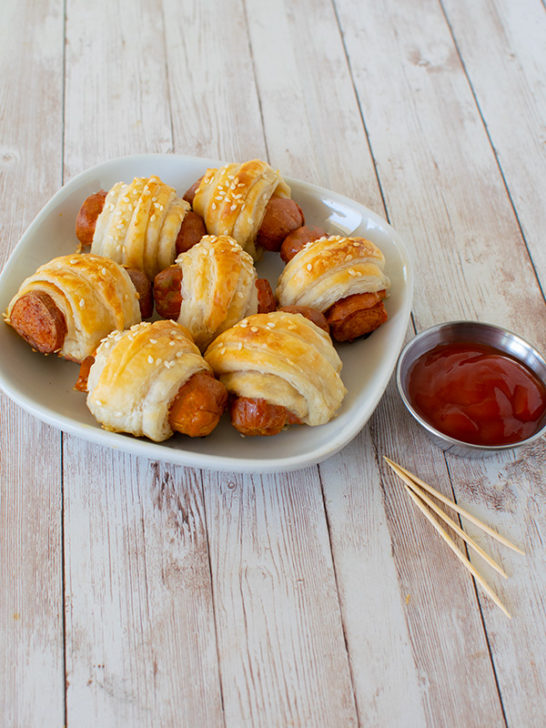 Kosher Franks in Blankets on a square white plate next to a small metal bowl with ketchup and a few toothpicks all on a white slatted wood table