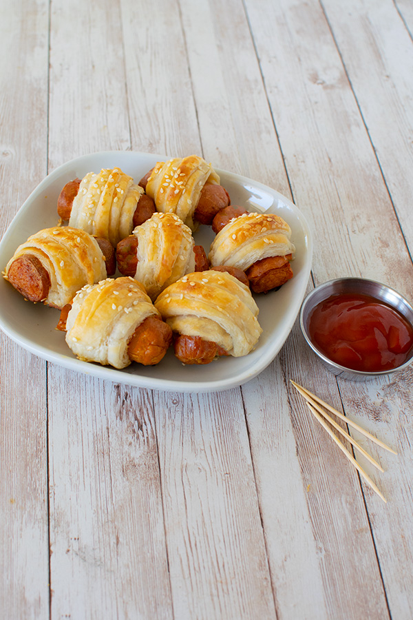 Kosher Franks in Blankets on a square white plate next to a small metal bowl with ketchup and a few toothpicks all on a white slatted wood table