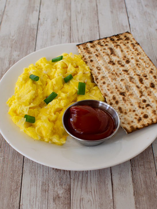Diary free scrambled eggs on a white plate with a piece of matzo and a little metal bowl of ketchup on a white wood table