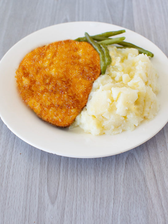 Cornflake fried chicken on a white plate with mashed potatoes and green beans on a light gray wood table.