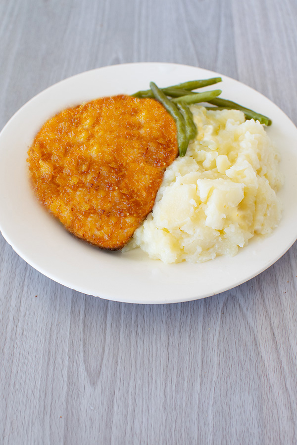Cornflake fried chicken on a white plate with mashed potatoes and green beans on a light gray wood table.