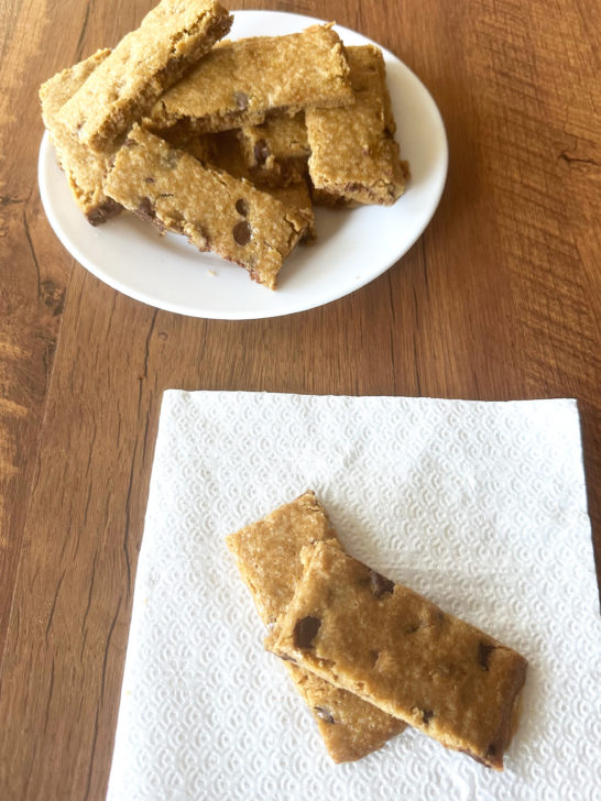 Two Easy chocolate chip cookie bars on a napkin with more bars on a white plate in the background, all on a dark wood table.