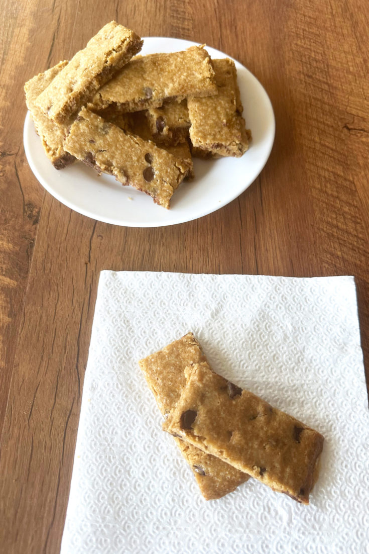 Two Easy chocolate chip cookie bars on a napkin with more bars on a white plate in the background, all on a dark wood table.