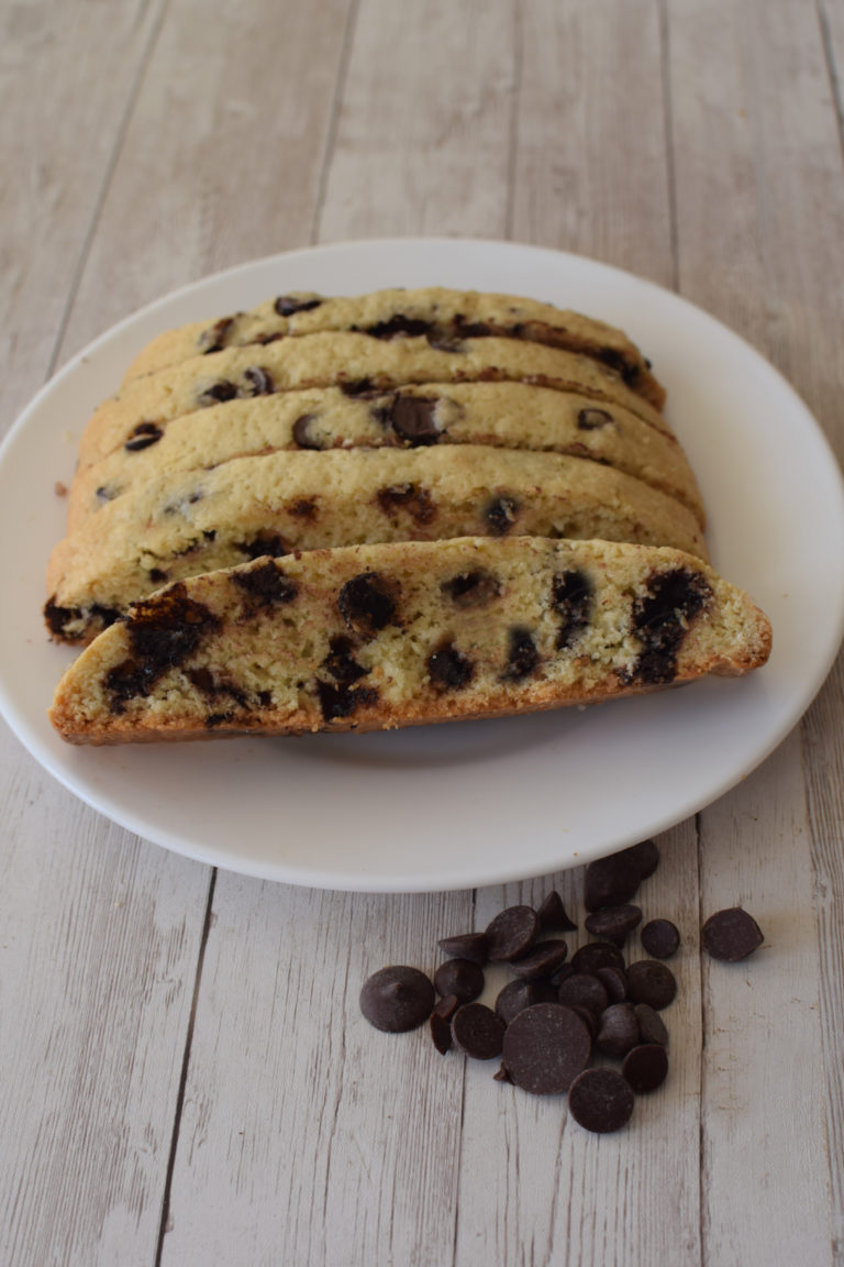 Slices of Chocolate Chip Mandel Bread on a white plate on a white wooden table with chocolate chips nearby