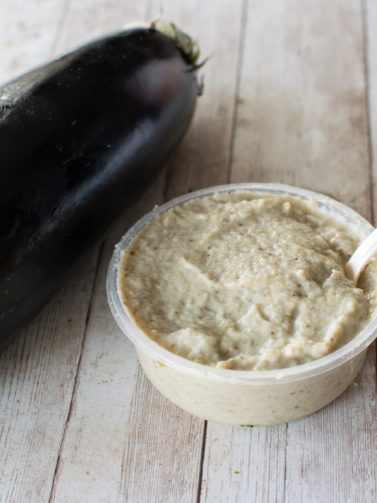 Eggplant salad with tahini in a container with a spoon near a raw eggplant on a white wood table.
