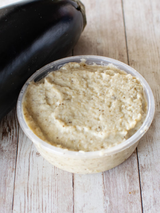 Eggplant salad with mayonnaise in a container with a spoon near a raw eggplant, all on a white wood table.