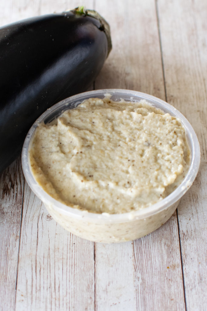 Eggplant salad with mayonnaise in a container with a spoon and an eggplant on a white wood table