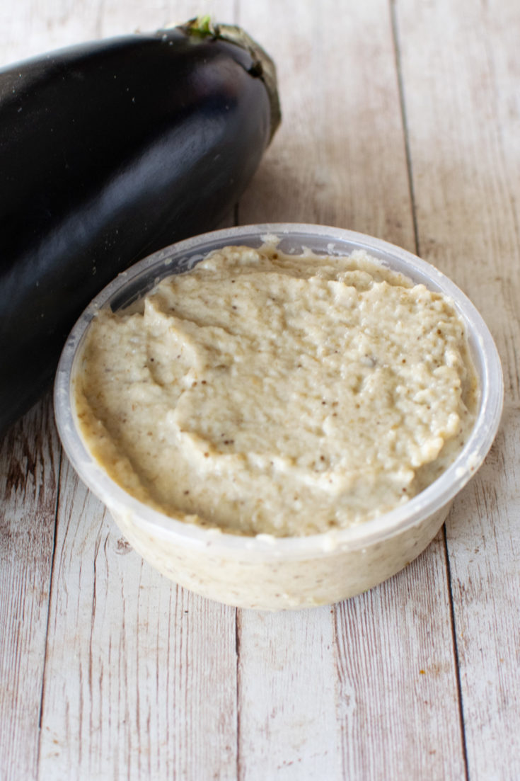 Eggplant salad with mayonnaise in a container with a spoon near a raw eggplant, all on a white wood table.