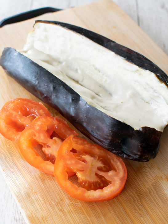 Roasted eggplant sliced open with tehini poured on near slices of tomato, all on a wooden cutting board.