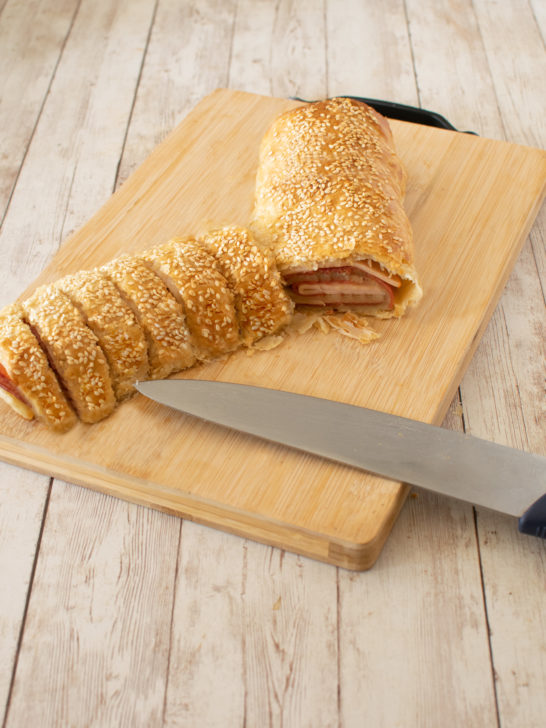 Deli roll with slices on a wooden cutting board and a knife on a white wood table