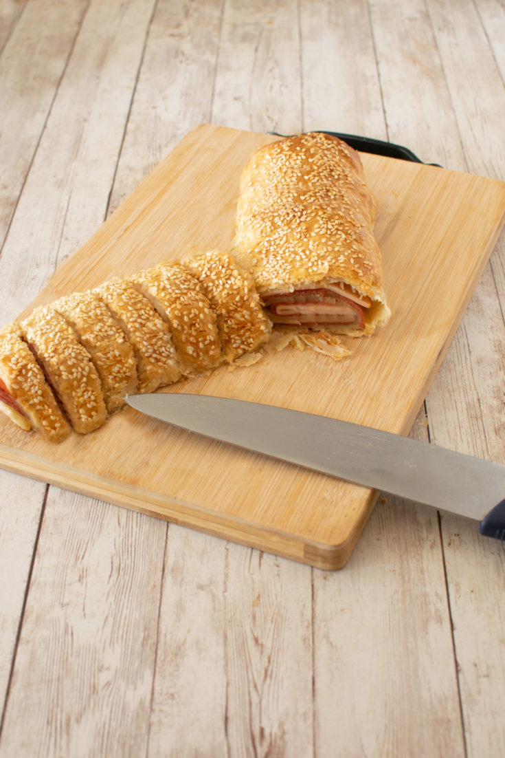 Deli roll with slices on a wooden cutting board and a knife on a white wood table