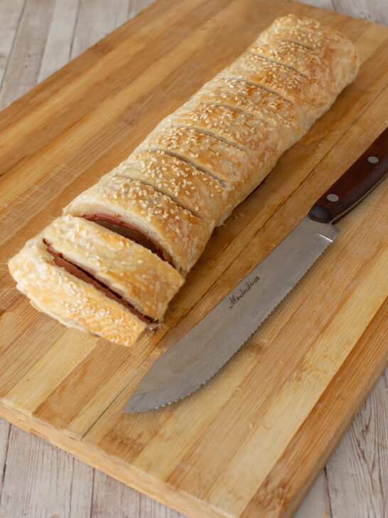 Sliced Kosher deli roll on a wooden cutting board next to a knife on a white wood table.