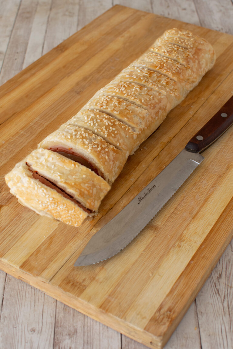 Sliced Kosher deli roll on a wooden cutting board next to a knife on a white wood table.