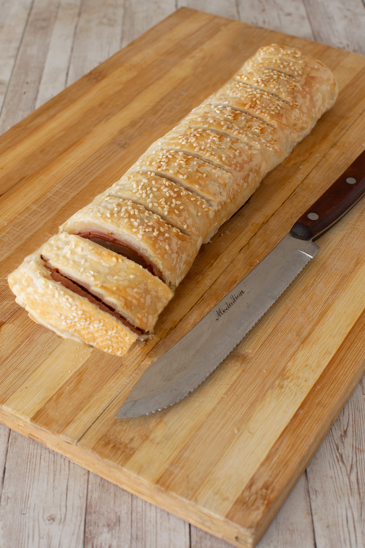 Sliced Kosher deli roll on a wooden cutting board next to a knife on a white wood table.