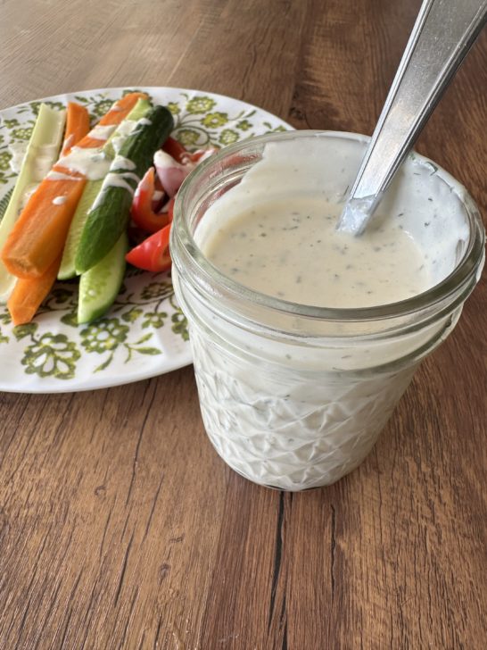 A jar of Dairy-Free Homemade Ranch Dressing with a spoon on a dark wood table next to a plate with cut raw vegetables.