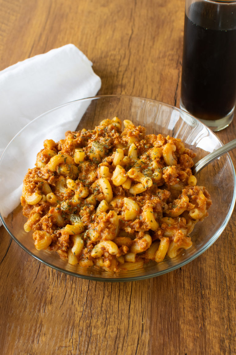 Easy American Goulash in a clear bowl with a fork on a dark wood table with a white napkin to the left and a clear glass of coke on the right