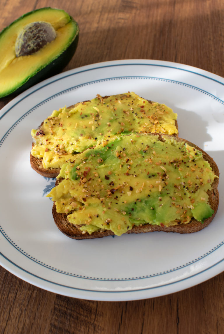 2 slices of Really easy avocado toast on a white place with half and avocado nearby on a dark wood table.