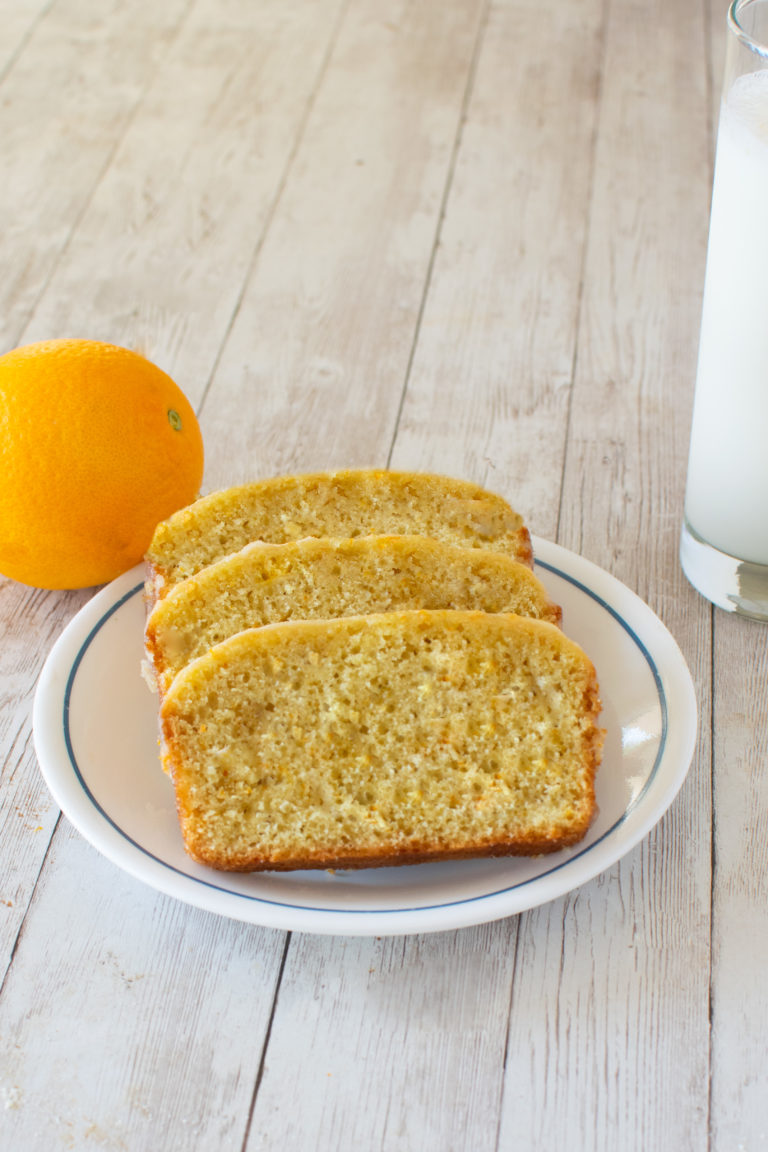 Easy Dairy-Free Orange Loaf Cake slices on a white plate with an orange and a clear glass of milk nearby on a white wood table