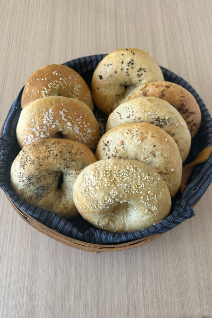 Bagels with a variety of toppings in a lined wicker basket on a white wood table.
