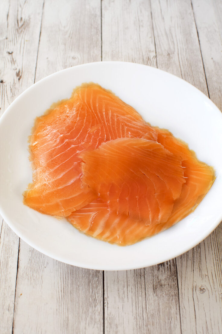 A few pieces of homemade dill-cured lox on a white plate on a white wood table