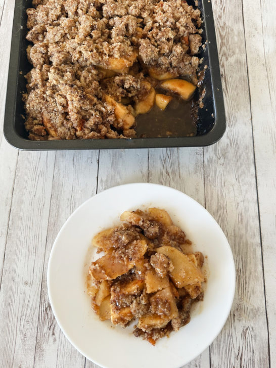 Dairy-free apple crumble on a white plate with a pan of the apple crumble in the background, all on a white wood table.